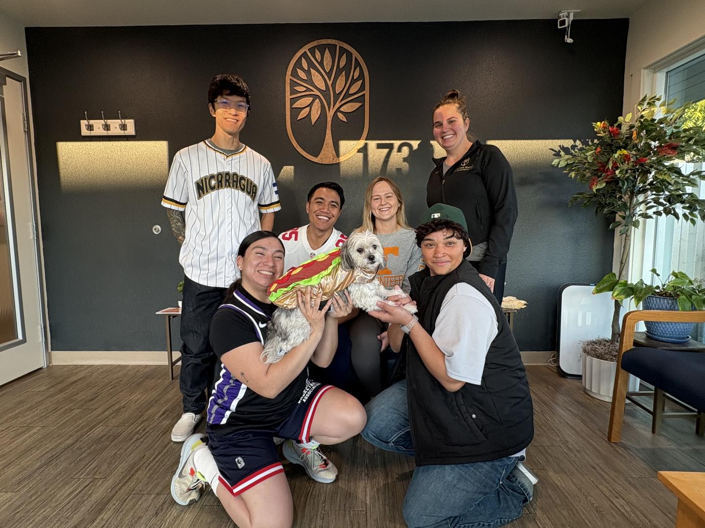 Dr. Tex Mabalon's team in casual jerseys and hats, kneeling around their therapy dog, Dr. Pepper, who is wearing a hot dog costume.