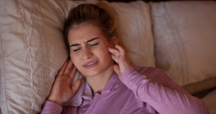 A woman lying in bed holding her jaw in discomfort, representing sudden TMJ symptoms often caused by overnight teeth clenching or bruxism.
