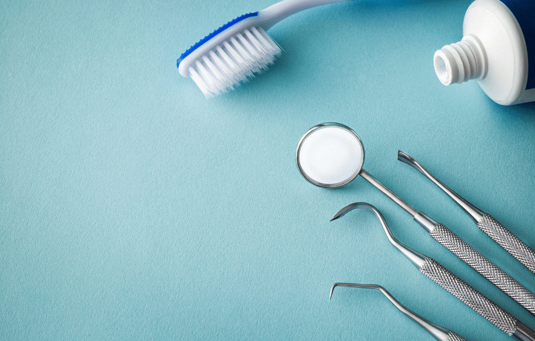 A top-down view of dental professional tools including a mirror, explorer, and scaler next to a toothbrush and tube of toothpaste on a blue surface.