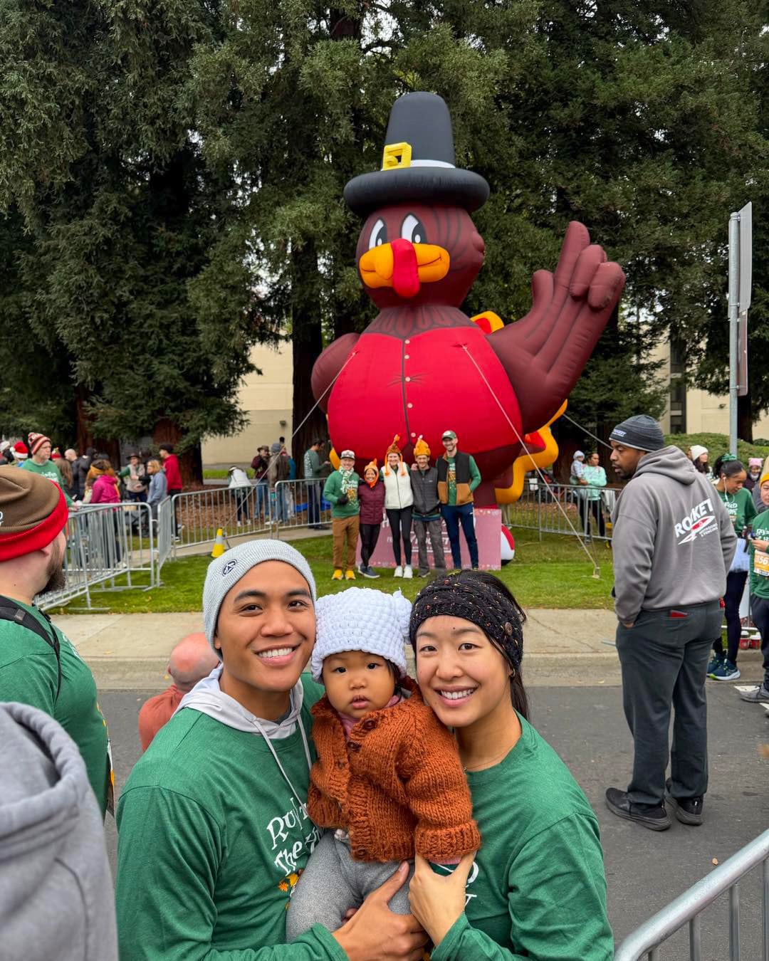 A happy family of three smiling at a Sacramento community event with a large inflatable turkey in the background.